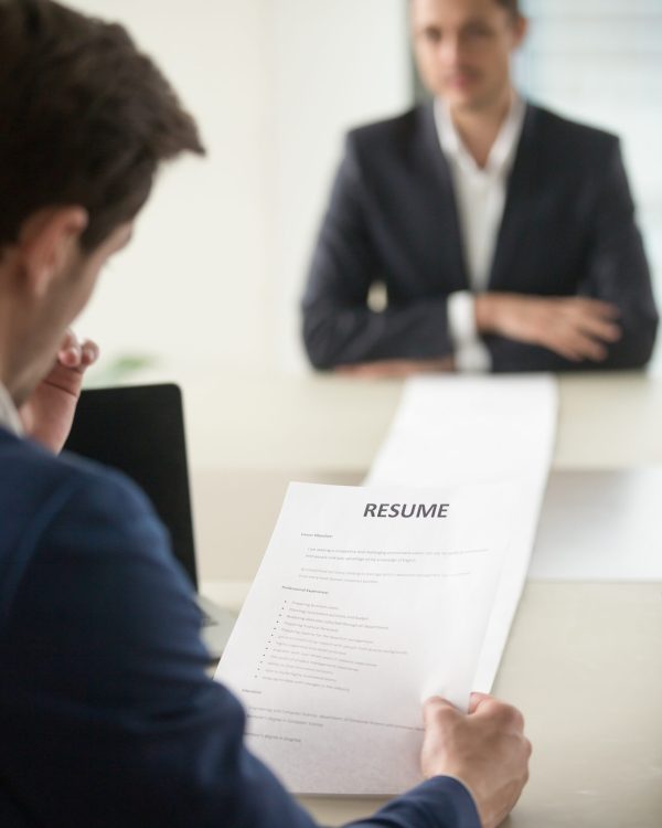 Back view of employer reading resume focus on document, job applicant waiting for hiring decision at desk on background. Hiring manager studying long list of candidates achievements and experiences